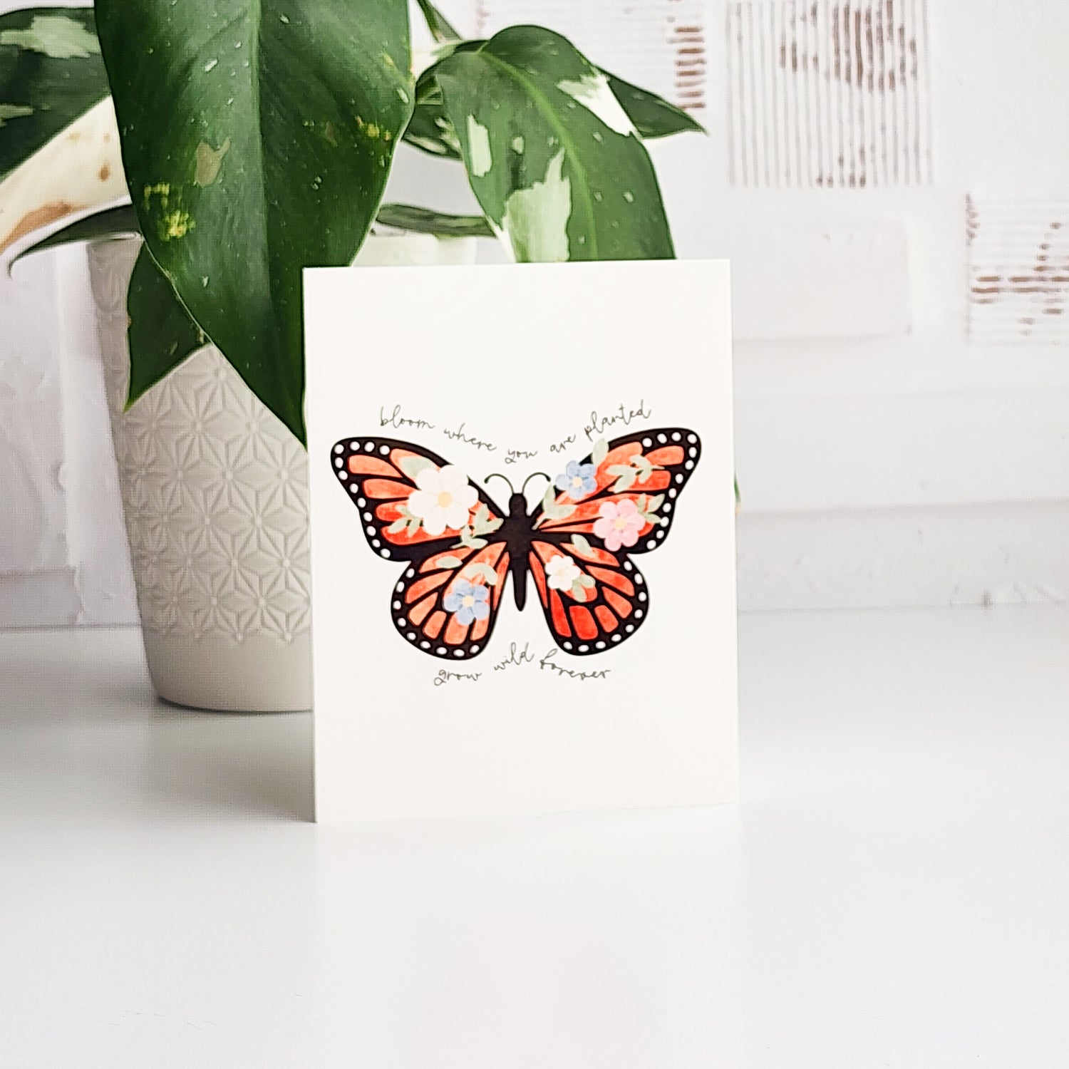 greeting card with an illustrated butterfly and the words bloom where you are planted, grow wild forever. The card is on a white background next to a potted plant