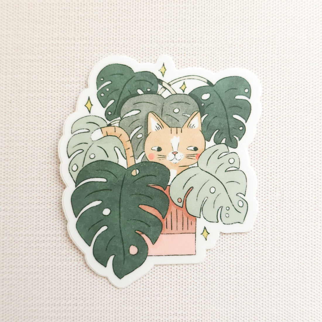 sticker of a cat in a monstera plant on a linen background