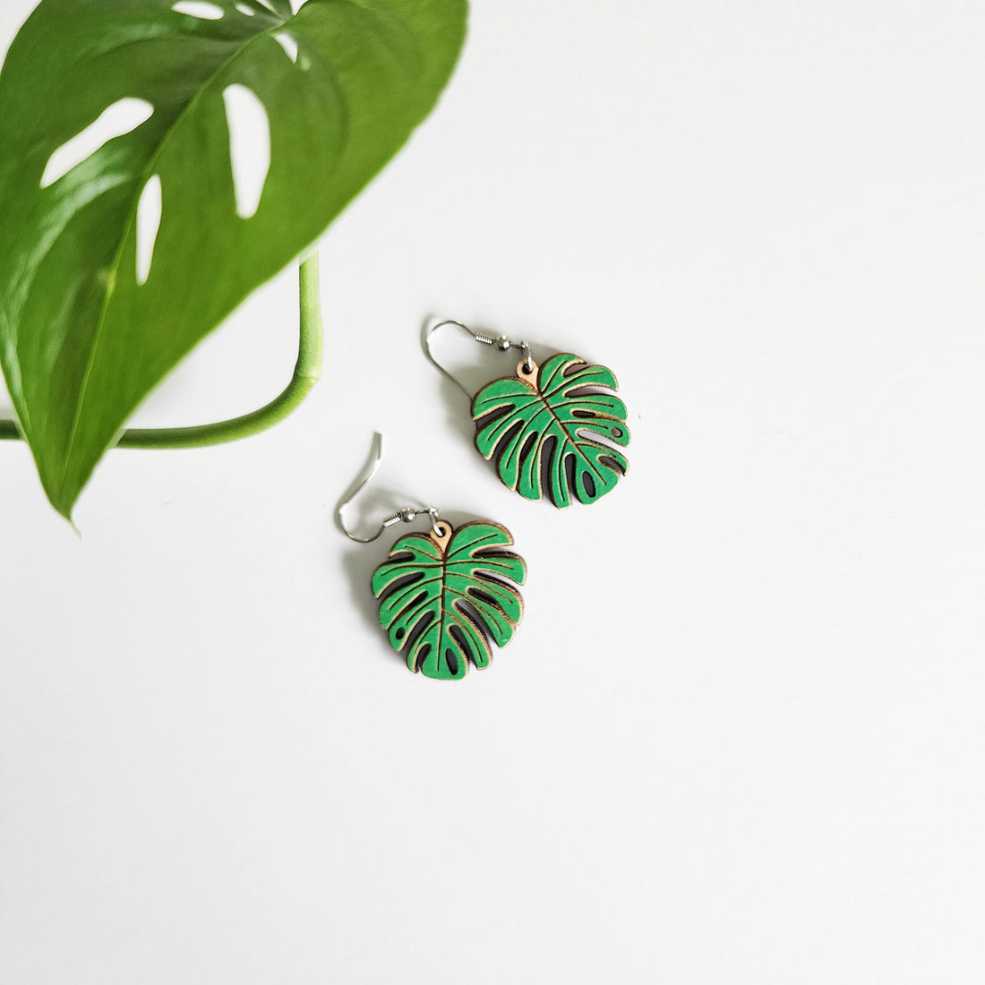pair of monstera leaf earrings on a white background next to a leaf