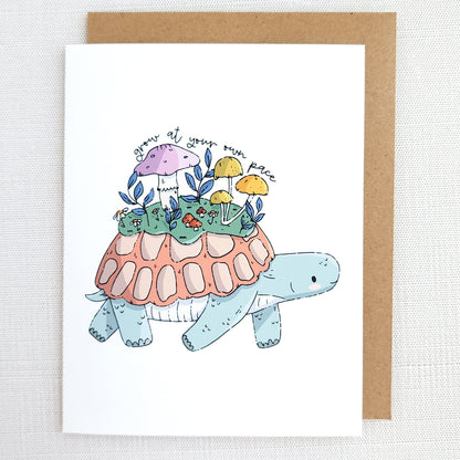 Greeting card with a turtle illustration carrying plants and mushrooms on its shell and the words grow at your own pace