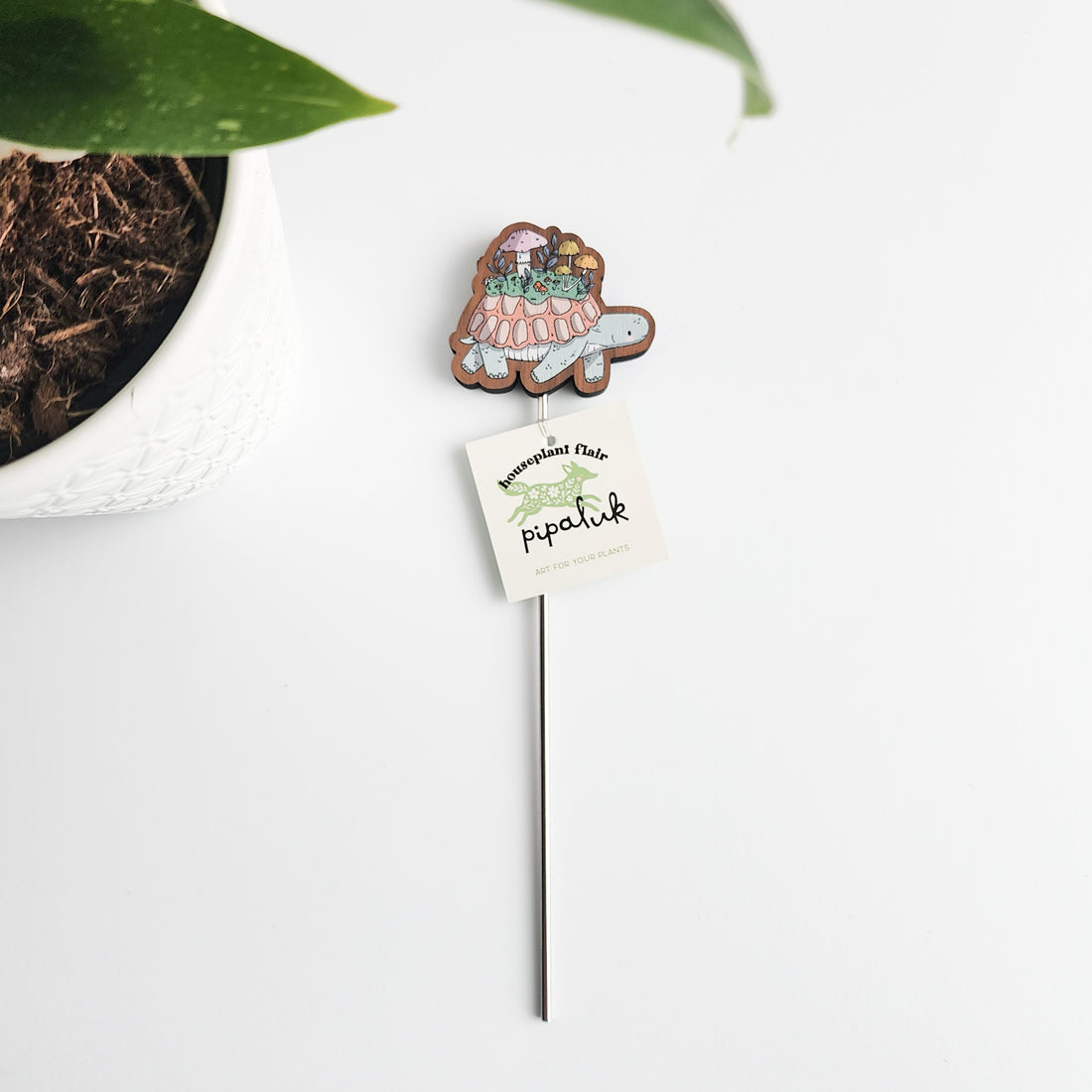 turtle plant pick with a pipaluk tag on a white background next to a plant