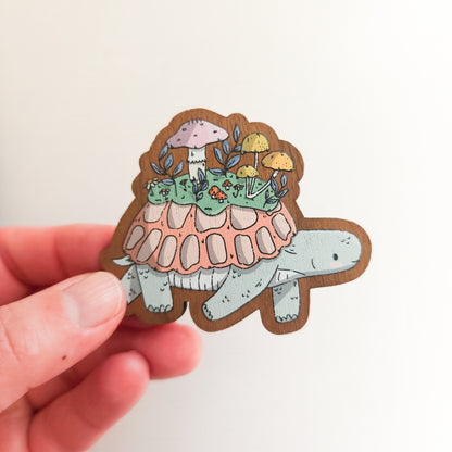 hand holding a wooden magnet with an illustration of a turtle with a mushroom meadow on its back on a linen background