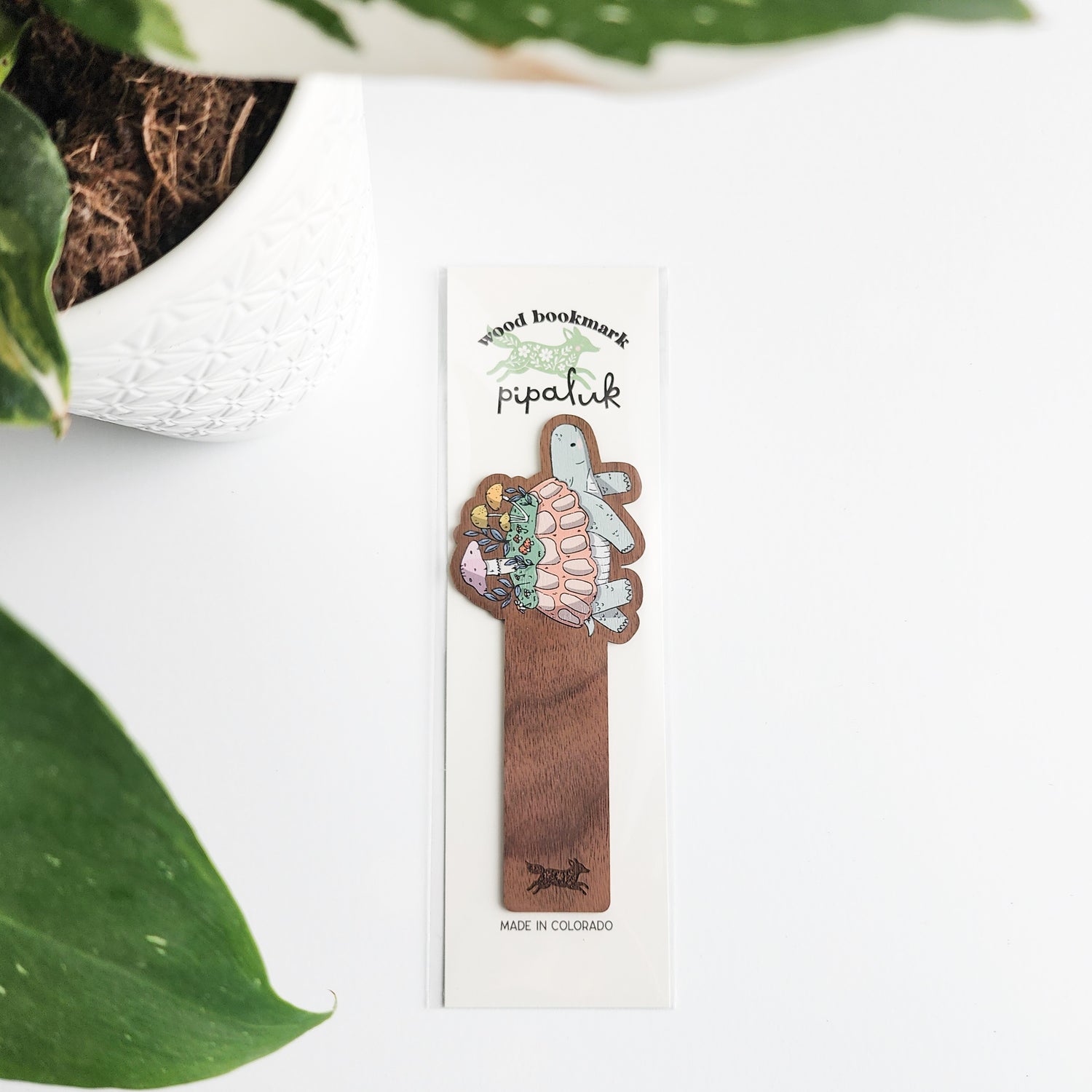 Wooden bookmark with turtle design in pipaluk packaging, surrounded by green leaves.