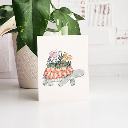illustrated card with a turtle growing a meadow on its back and the words grow at your own pace. The card is on a white background next to a plant