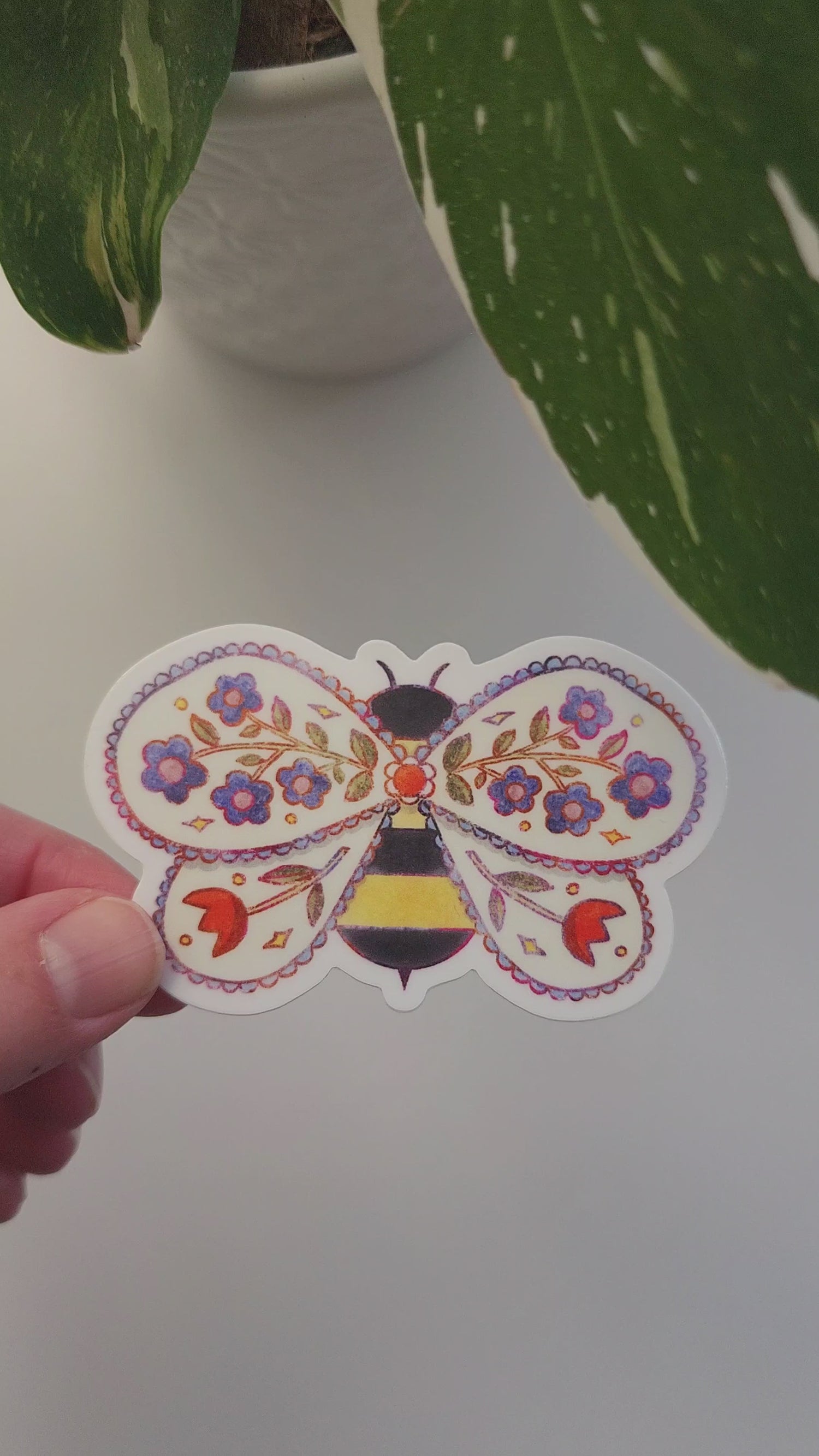 video of floral bee sticker held in a hand next to a potted plant