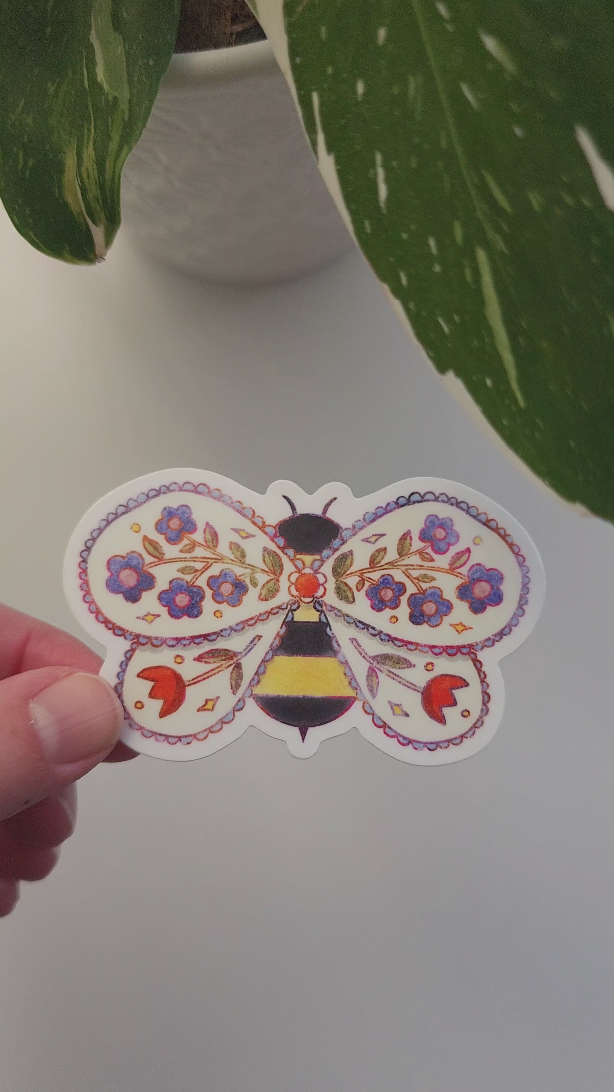 video of floral bee sticker held in a hand next to a potted plant