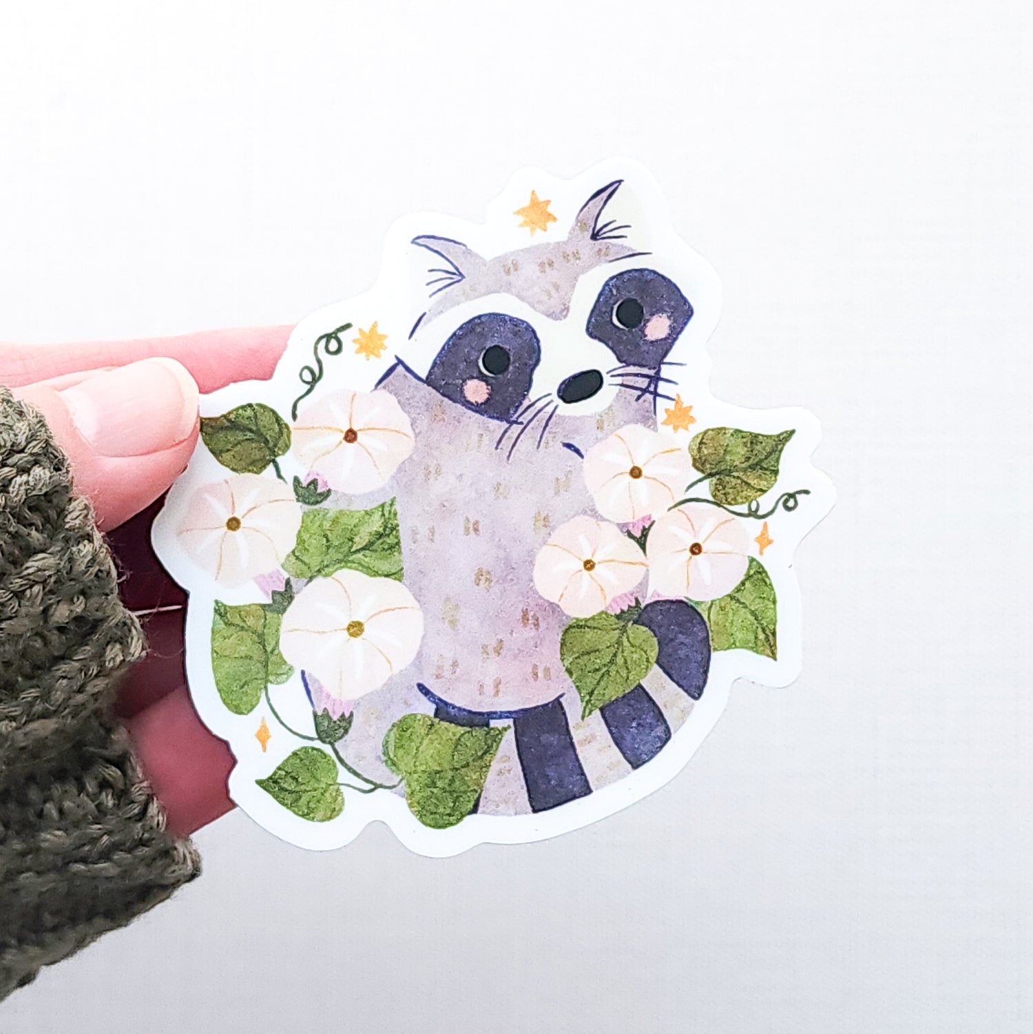 Vinyl sticker featuring a curious raccoon with dark eye markings, surrounded by white moonflowers, green vines, and tiny golden stars, held against a light background.