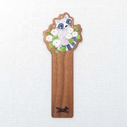 Wooden bookmark featuring a curious raccoon surrounded by white moonflowers, green leaves, and tiny golden stars, printed on walnut wood and photographed on a neutral fabric background.