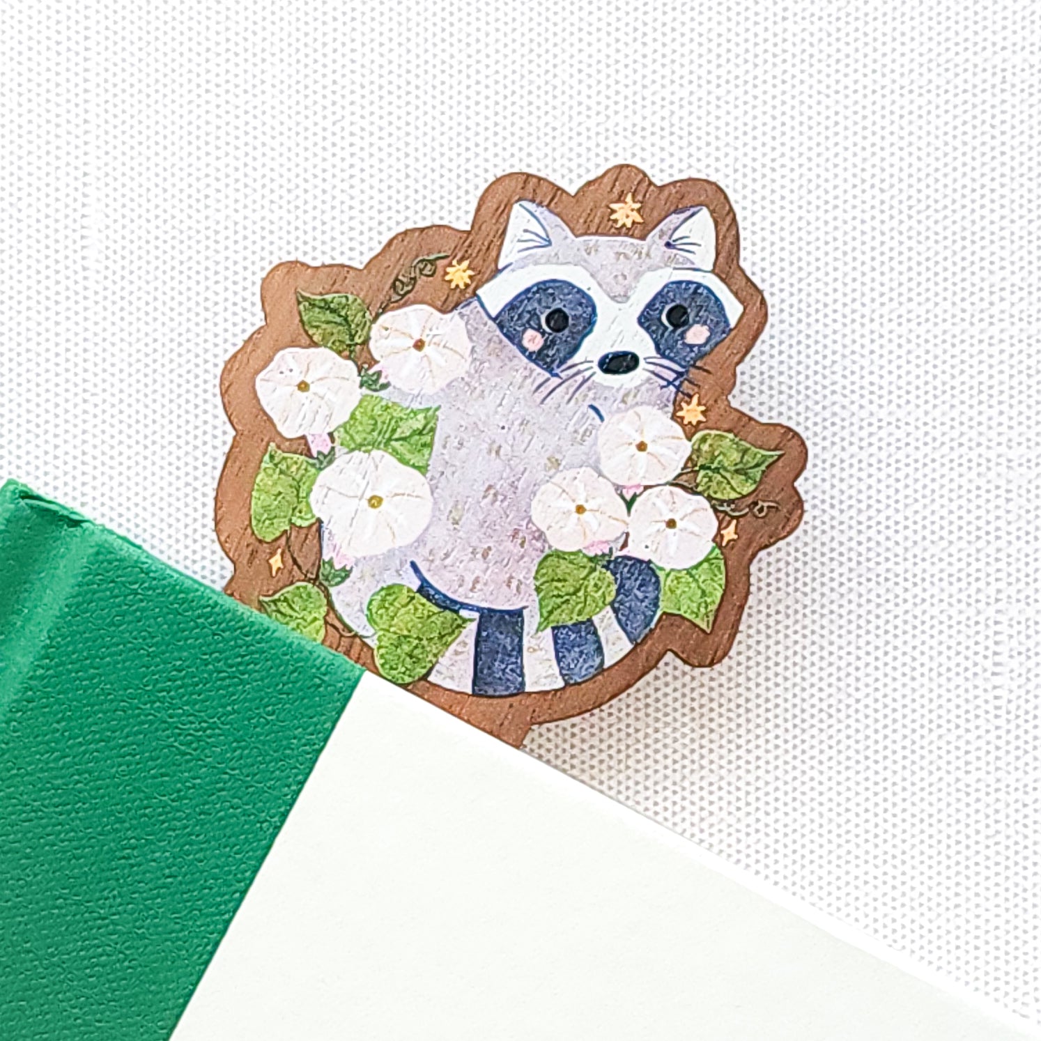 Wooden bookmark featuring a curious raccoon surrounded by white moonflowers, green leaves, and tiny golden stars, printed on walnut wood and photographed in a book on a neutral fabric background.