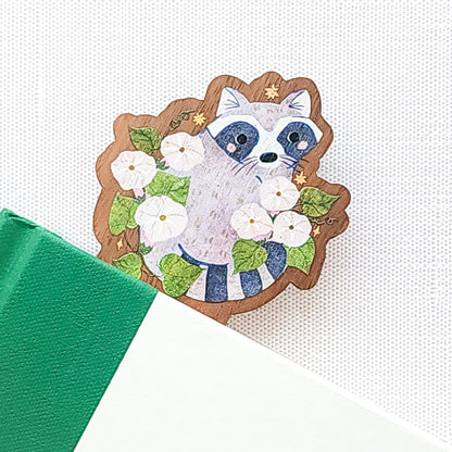Wooden bookmark featuring a curious raccoon surrounded by white moonflowers, green leaves, and tiny golden stars, printed on walnut wood and photographed in a book on a neutral fabric background.