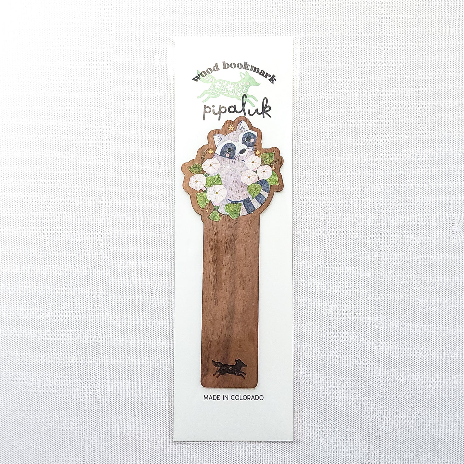 Wooden bookmark featuring a curious raccoon surrounded by white moonflowers, green leaves, and tiny golden stars, printed on walnut wood and photographed in Pipaluk branded packaging on a neutral fabric background.