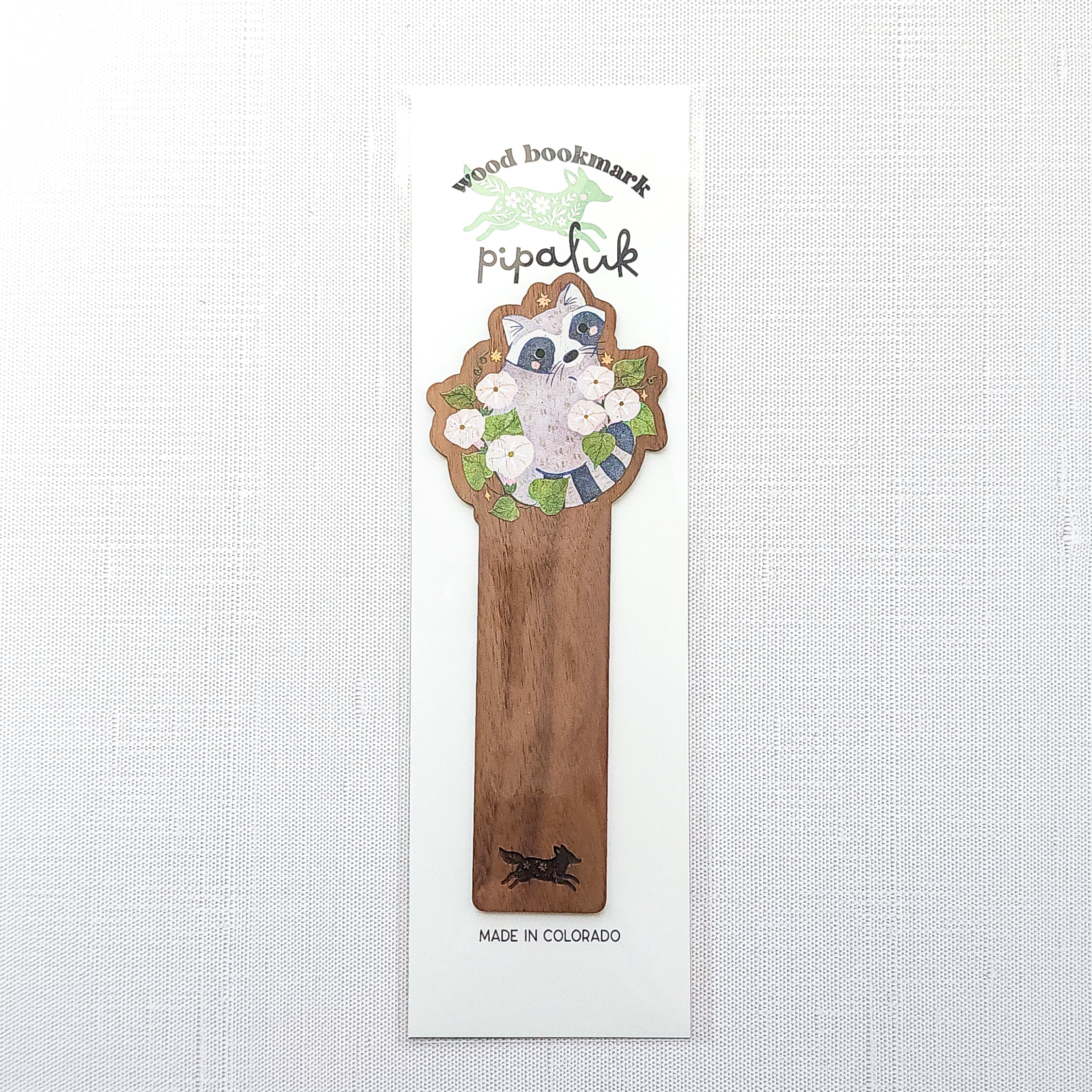 Wooden bookmark featuring a curious raccoon surrounded by white moonflowers, green leaves, and tiny golden stars, printed on walnut wood and photographed in Pipaluk branded packaging on a neutral fabric background.