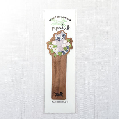Wooden bookmark featuring a curious raccoon surrounded by white moonflowers, green leaves, and tiny golden stars, printed on walnut wood and photographed in Pipaluk branded packaging on a neutral fabric background.