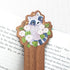Wooden bookmark featuring a curious raccoon surrounded by white moonflowers, green leaves, and tiny golden stars, printed on walnut wood and photographed on a book on a neutral fabric background.