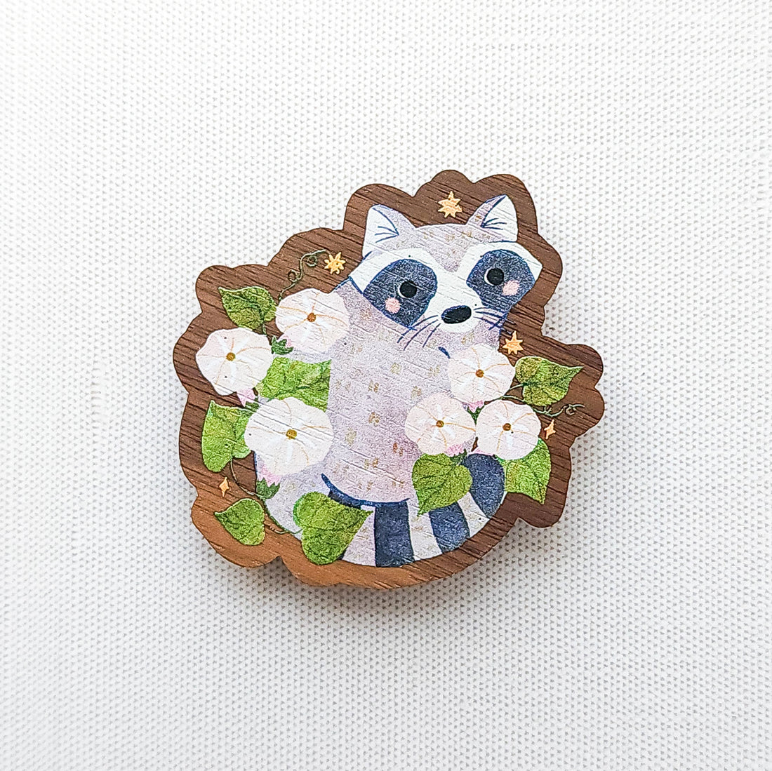 Wooden magnet featuring a curious raccoon surrounded by white moonflowers, green leaves, and tiny golden stars, printed on walnut wood and photographed on a neutral fabric background.
