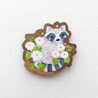 Wooden magnet featuring a curious raccoon surrounded by white moonflowers, green leaves, and tiny golden stars, printed on walnut wood and photographed on a neutral fabric background.