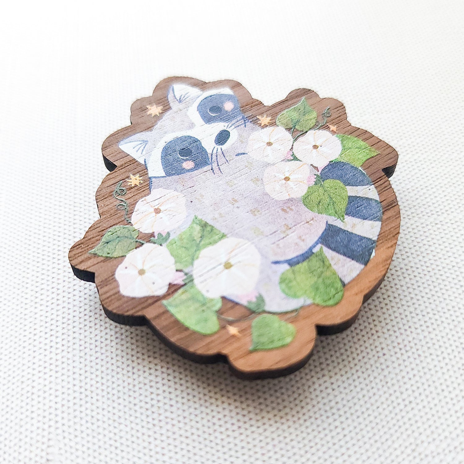 Wooden magnet featuring a curious raccoon surrounded by white moonflowers, green leaves, and tiny golden stars, printed on walnut wood and photographed on a neutral fabric background.