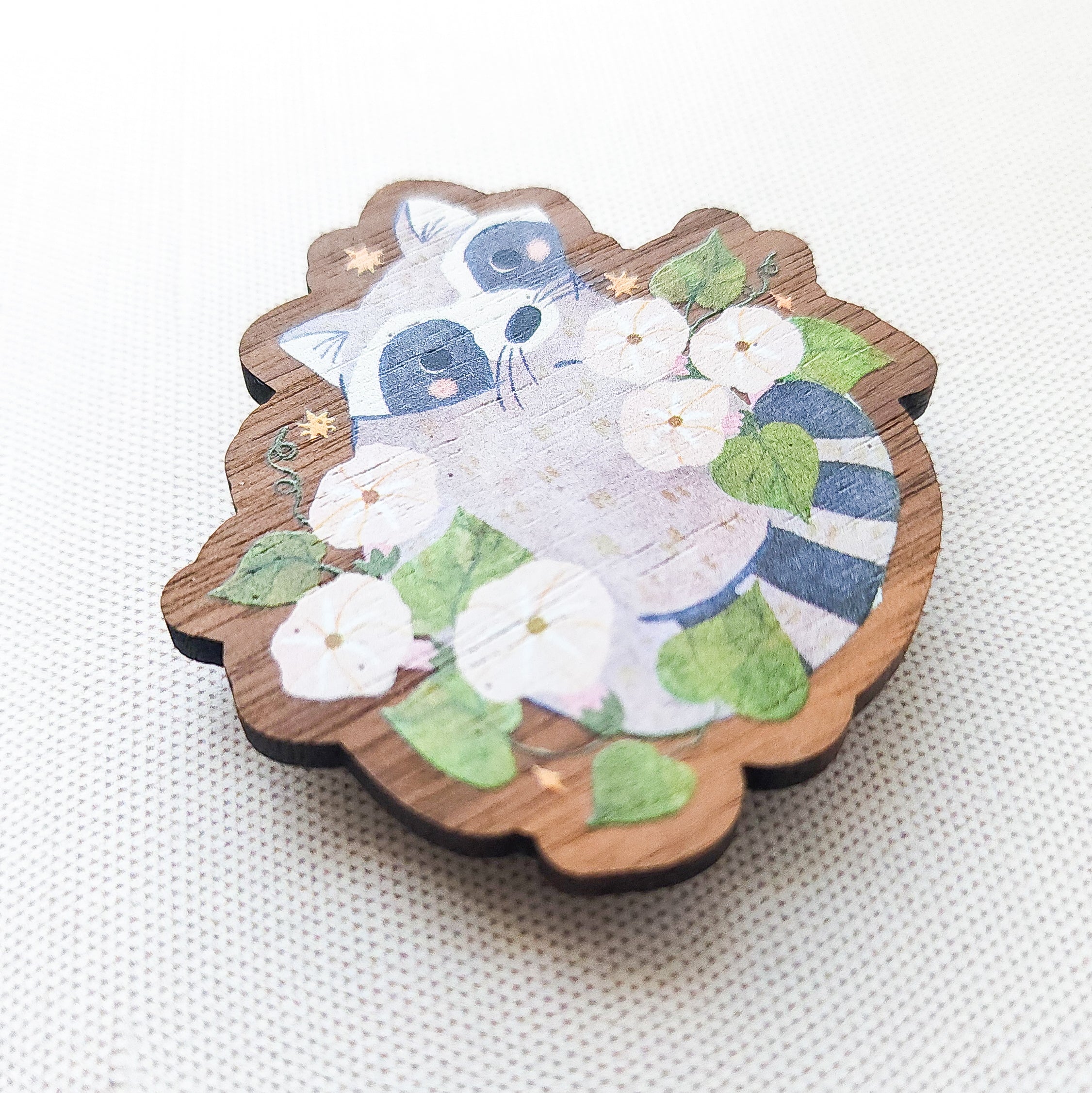 Wooden magnet featuring a curious raccoon surrounded by white moonflowers, green leaves, and tiny golden stars, printed on walnut wood and photographed on a neutral fabric background.