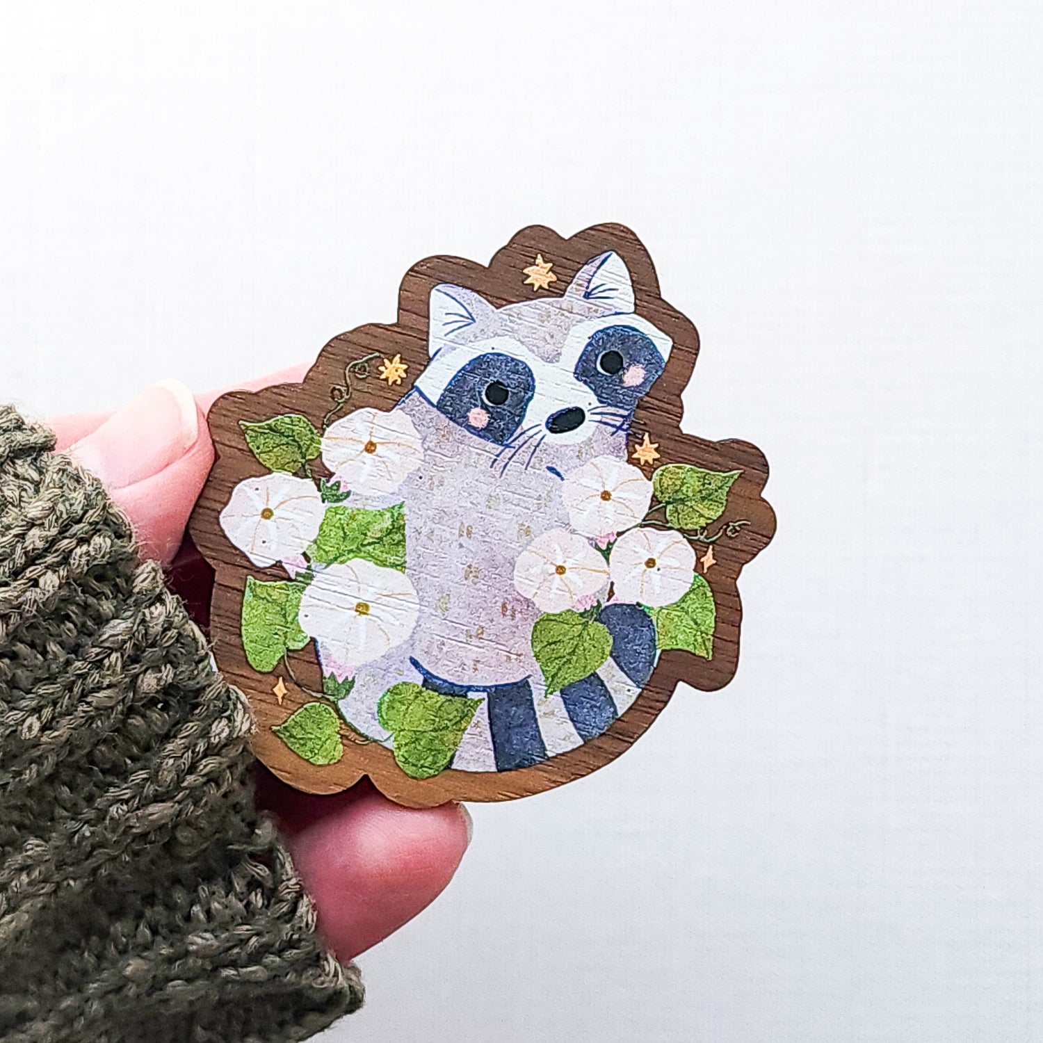 Wooden magnet featuring a curious raccoon surrounded by white moonflowers, green leaves, and tiny golden stars, printed on walnut wood and photographed held in a hand on a neutral fabric background.