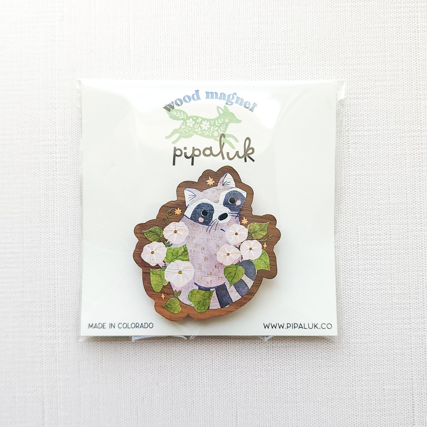 Wooden magnet featuring a curious raccoon surrounded by white moonflowers, green leaves, and tiny golden stars, printed on walnut wood and photographed in pipaluk packaging on a neutral fabric background.