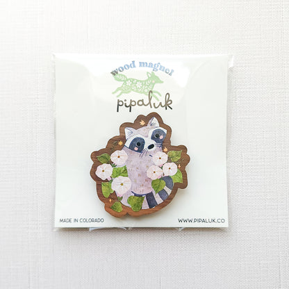 Wooden magnet featuring a curious raccoon surrounded by white moonflowers, green leaves, and tiny golden stars, printed on walnut wood and photographed in pipaluk packaging on a neutral fabric background.