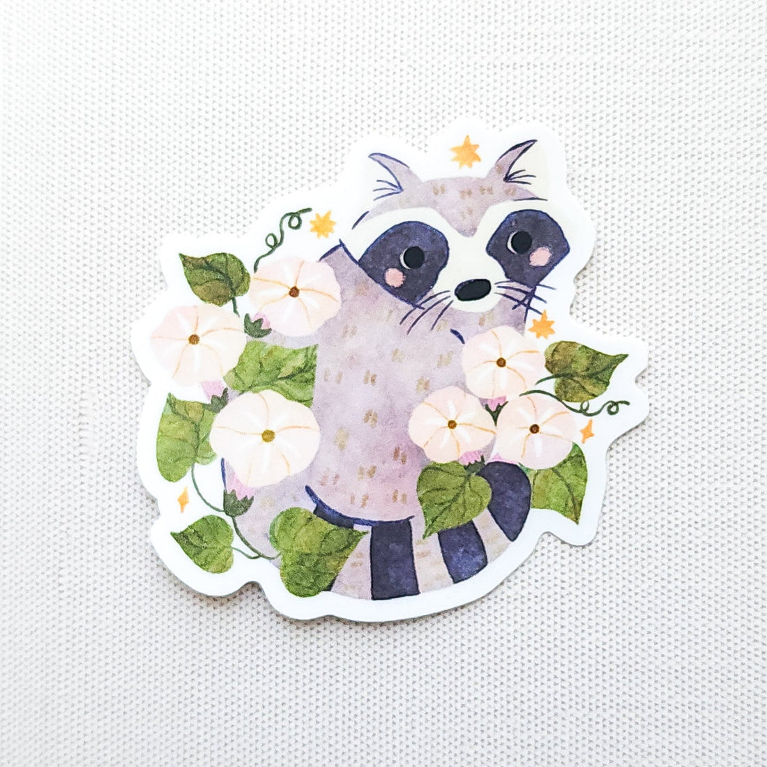 Vinyl sticker featuring a curious raccoon with dark eye markings, surrounded by white moonflowers, green vines, and tiny golden stars photographed on a neutral background
