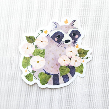 Vinyl sticker featuring a curious raccoon with dark eye markings, surrounded by white moonflowers, green vines, and tiny golden stars photographed on a neutral background