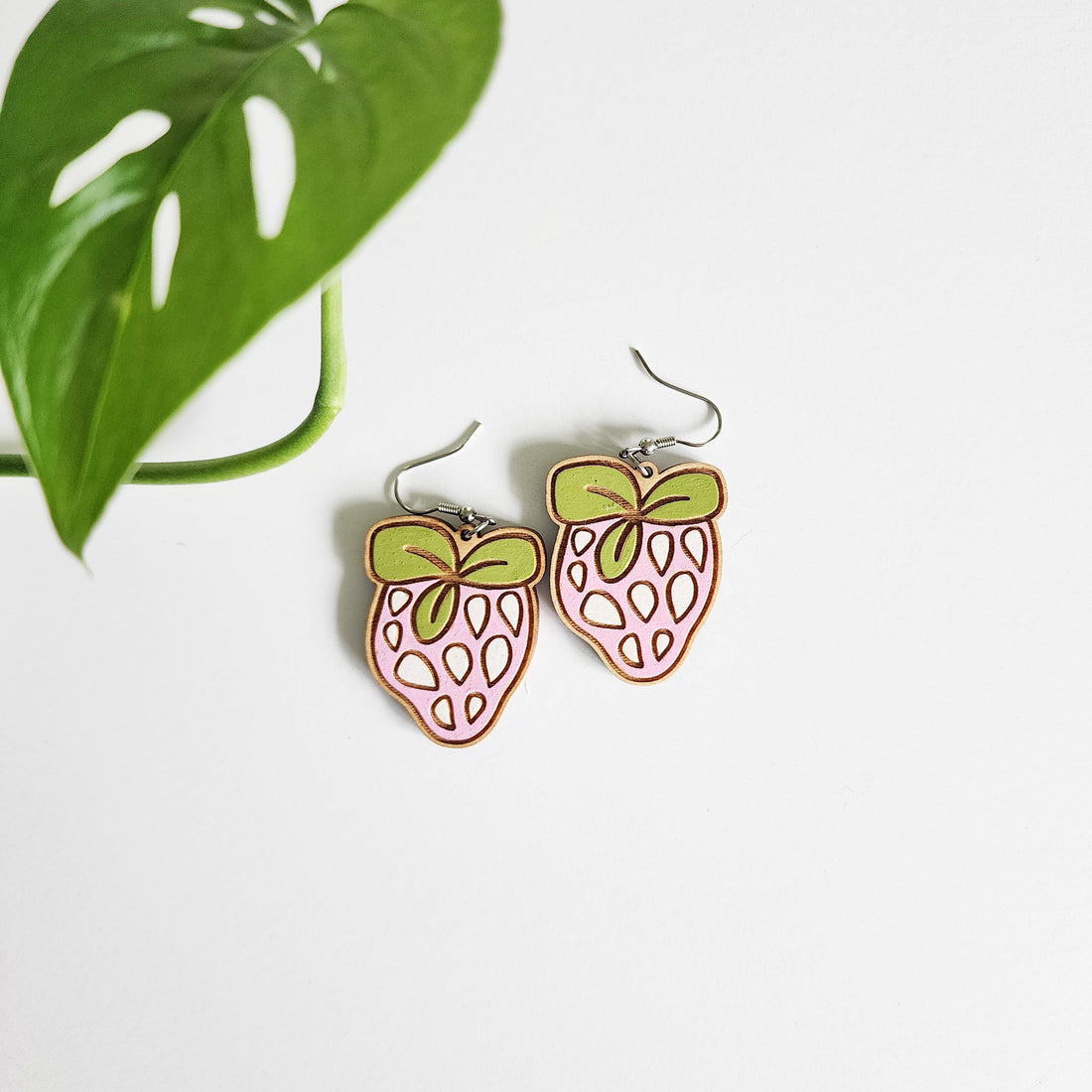 pair of strawberry earrings on white background next to a leaf