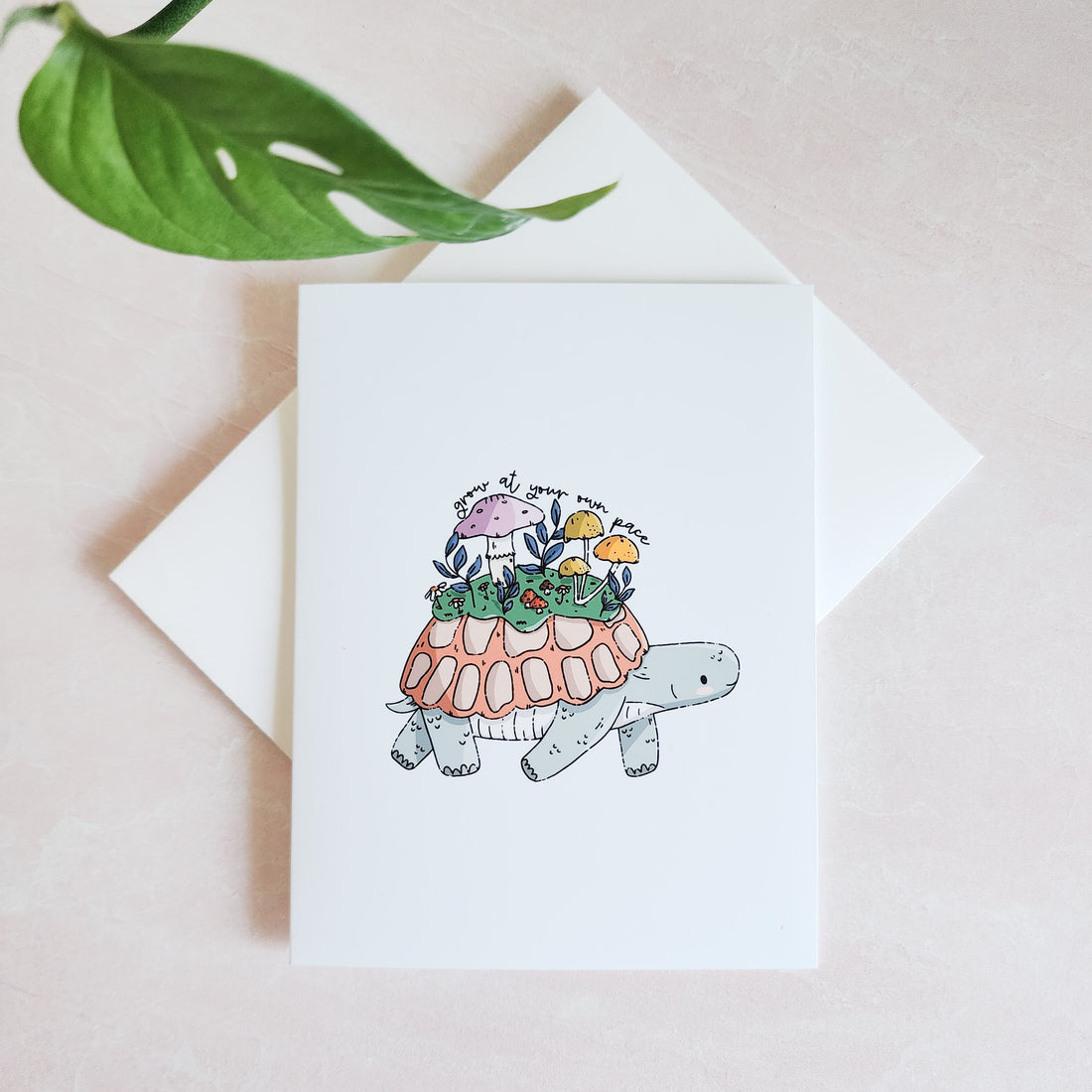 greeting card depicting a turtle with mushrooms on its back on top of a white envelope